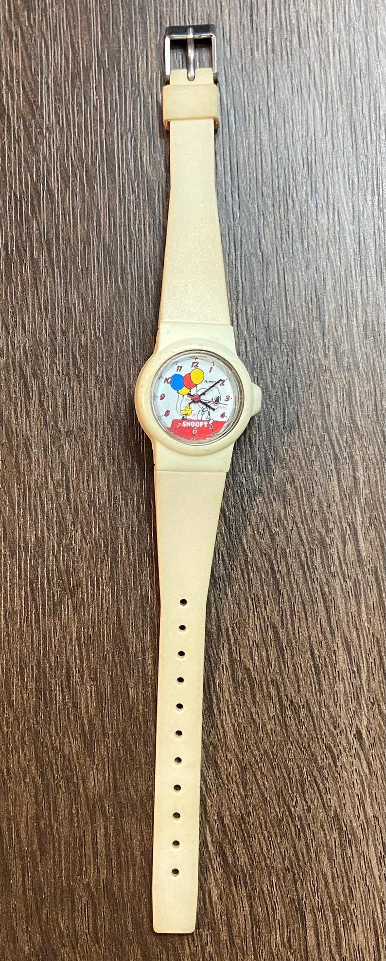 May include: Cream-colored Snoopy wristwatch with a matching band. The watch face features Snoopy holding balloons. The watch band has a buckle closure and multiple adjustment holes. A classic, vintage accessory.
