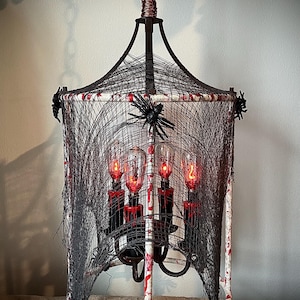 May include: A Halloween-themed chandelier with a black web design. The white frame is decorated with red splatters, and black spiders are attached. The chandelier has four red-lit bulbs, creating a spooky atmosphere.
