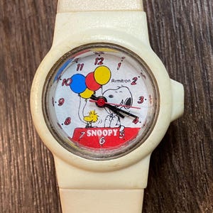 May include: Cream-colored Armitron watch featuring Snoopy and Woodstock with balloons. The watch face has a red, white, and blue color scheme with the word "Snoopy" in red. The watch band is also cream-colored.