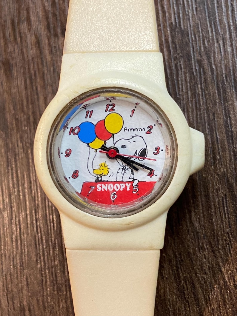 May include: Cream-colored Armitron watch featuring Snoopy and Woodstock with balloons. The watch face has a red, white, and blue color scheme with the word "Snoopy" in red. The watch band is also cream-colored.