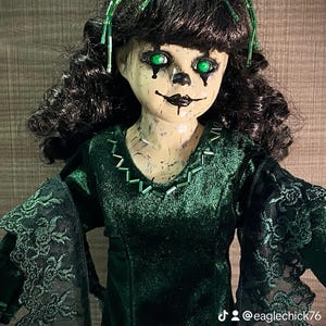 May include: A creepy porcelain doll with green eyes and a dark green velvet dress. The doll is wearing a crown of green beads and has a painted-on smile.