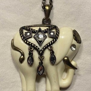 May include: A white elephant pendant necklace with a gold-tone chain and decorative accents. The elephant has rhinestones on its body and a gold-tone trim around its head.