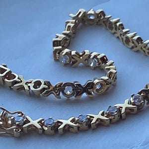 May include: A gold-toned bracelet featuring an alternating pattern of "X" and "O" shapes, each adorned with a small, clear gemstone. The bracelet is unclasped, showing the clasp mechanism.