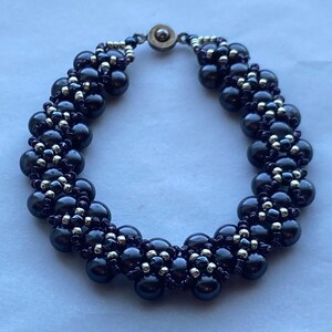 May include: A dark gray beaded bracelet with a silver clasp. The bracelet features large, round, dark gray beads interspersed with smaller silver beads. The beads are strung together in a woven pattern, creating a textured look.