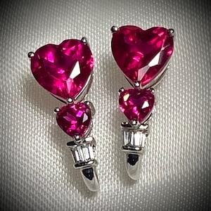 May include: A pair of silver earrings with three heart-shaped ruby gemstones. The earrings have a dangling design with a larger heart at the top and two smaller hearts below.
