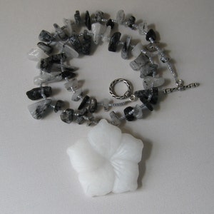 May include: A white flower pendant necklace with a silver clasp. The necklace is made of black and clear quartz stones.
