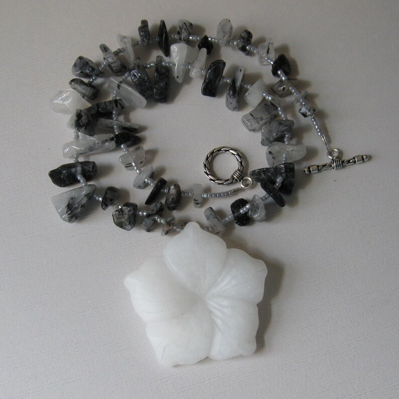 May include: A white flower pendant necklace with a silver clasp. The necklace is made of black and clear quartz stones.