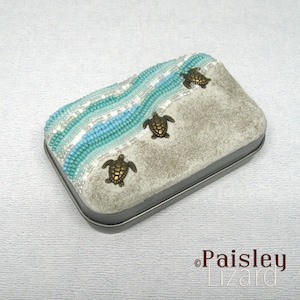 May include: A small, rectangular tin box decorated with a beaded design of turquoise and white waves.  Three small, gold-colored turtle charms are attached to the box.