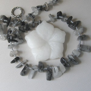 May include: A necklace made of black and white stones with a white flower pendant. The stones are faceted and have a natural, rough look. The necklace is strung on a silver chain.