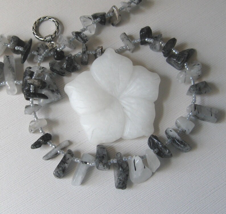 May include: A necklace made of black and white stones with a white flower pendant. The stones are faceted and have a natural, rough look. The necklace is strung on a silver chain.
