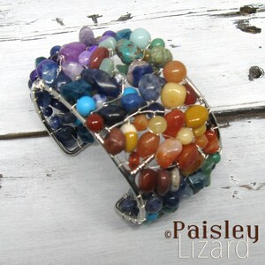 May include: A silver wire wrapped bracelet with a variety of colorful gemstones, including red, orange, yellow, green, blue, and purple. The bracelet is made with a wire mesh design and is a statement piece.