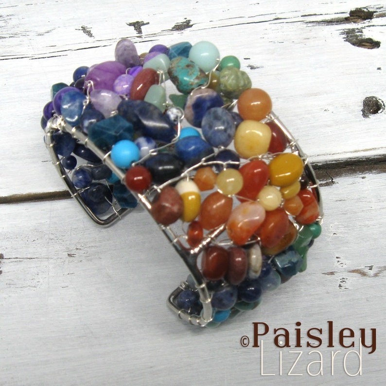 May include: A silver wire wrapped bracelet with a variety of colorful gemstones, including red, orange, yellow, green, blue, and purple. The bracelet is made with a wire mesh design and is a statement piece.