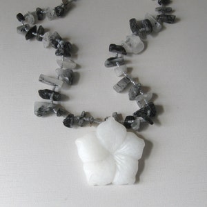 May include: A white carved flower pendant hangs from a necklace made of black and clear quartz stones.