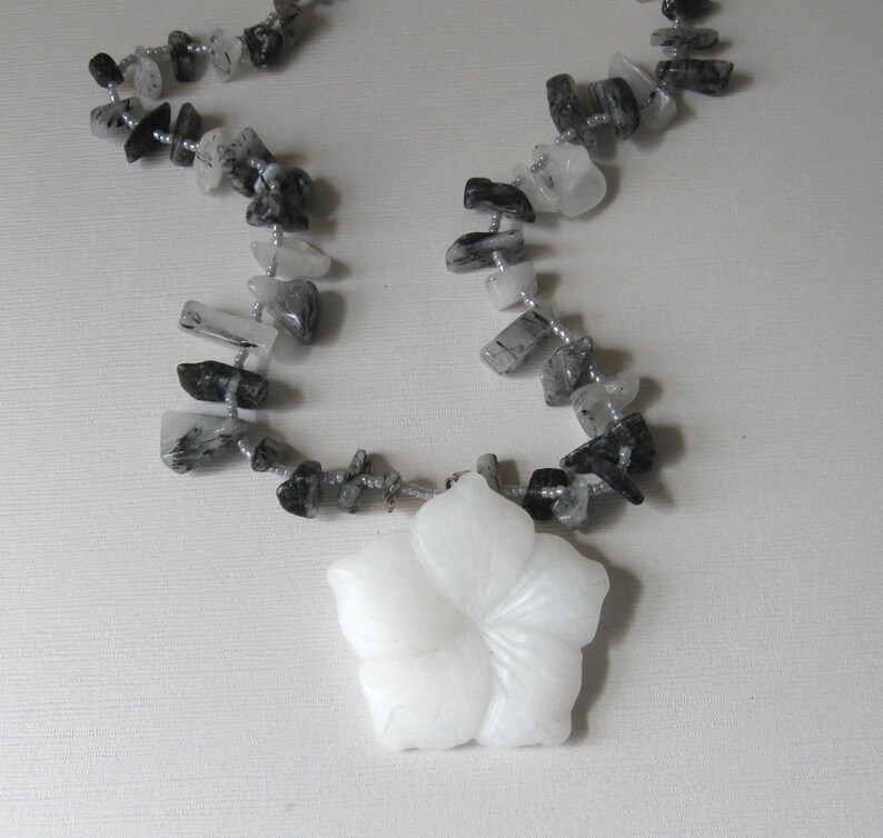 May include: A white carved flower pendant hangs from a necklace made of black and clear quartz stones.