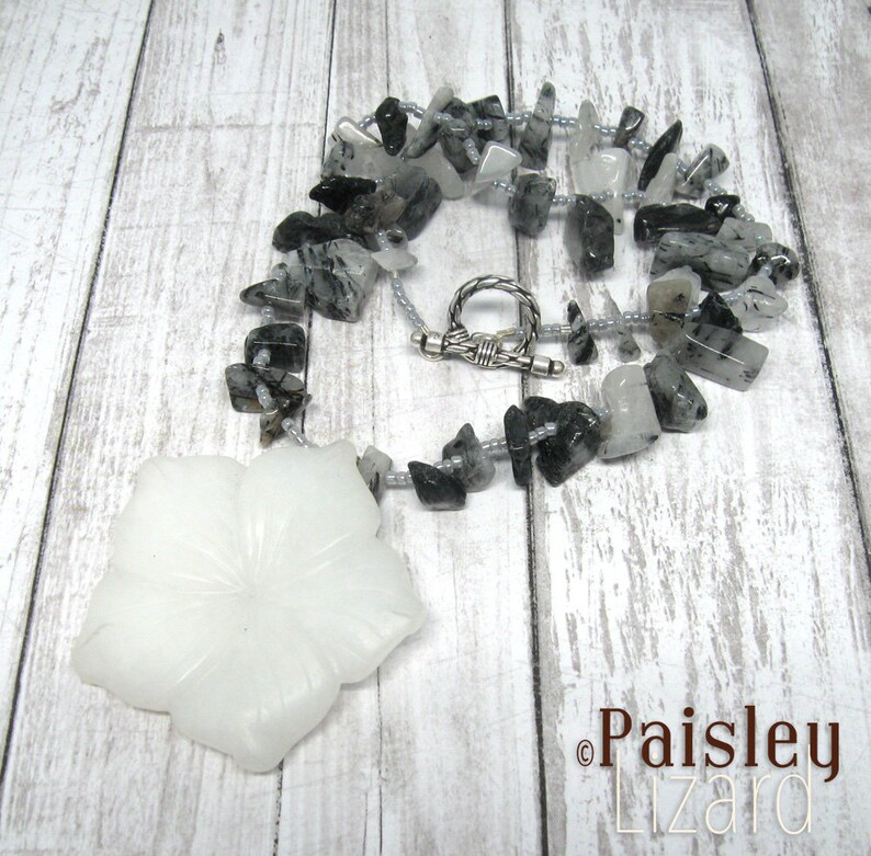 May include: A white flower pendant necklace with a silver clasp. The necklace is made of black and white stones and silver beads.