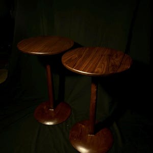 May include: Two matching side tables crafted from dark brown wood, each featuring a smooth, circular tabletop supported by a single central pedestal column that connects to a wide, flat, circular base, showcasing a rich, natural wood grain pattern.