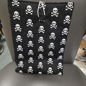 May include: Black fabric bag featuring a white skull and crossbones design. The bag has a button fastening and a white cord loop. The bag is on a grey chair.