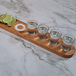 May include: Wooden tequila shot serving paddle with four shot glasses, lime wedges, and a salt ring. The paddle is made of light brown wood and has a handle. The shot glasses are filled with clear liquid.