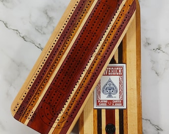 May include: Wooden cribbage board with a hinged design, featuring alternating stripes of light and dark wood. The board is open, revealing the pegging tracks and a deck of Maverick playing cards.