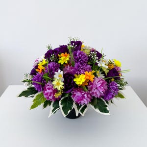 May include: A floral arrangement featuring deep purple roses, vibrant magenta pom-pom flowers, small yellow blossoms, and white daisy-like flowers. The bouquet is accented with green leaves that have white variegated edges, all set in a dark container.