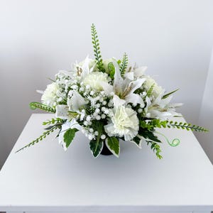 May include: A round floral arrangement composed of white lilies, carnations, and baby's breath. The arrangement is accented with green foliage and presented in a dark container. The flowers are arranged to create a full, abundant display.