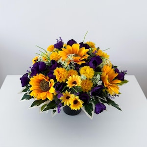 May include: A floral arrangement featuring bright yellow sunflowers and smaller yellow blossoms interspersed with deep purple flowers and green foliage. The flowers are contained within a dark, rounded base, creating a dense, rounded bouquet.
