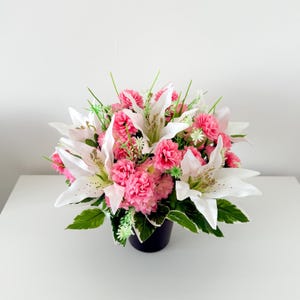May include: A colourful floral arrangement with white lilies, pink carnations, and green leaves. The flowers are arranged in a dark blue pot, creating a vibrant display. Ideal for home decor or as a gift, adding a touch of nature.