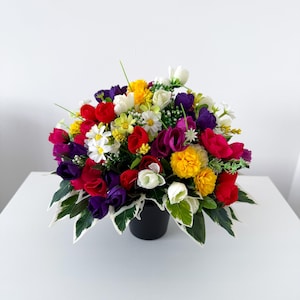 May include: A dense floral arrangement in a black pot featuring a mix of artificial flowers in vibrant shades of red, yellow, purple, and white. The bouquet includes various flower shapes and green leaves with white variegated edges throughout.