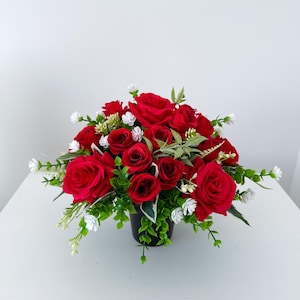 May include: A striking floral arrangement of red roses, complemented by small white flowers and green leaves. The arrangement is displayed in a black pot, creating a contrast against the white background. A classic decorative piece for the home.
