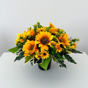 May include: A bright display of sunflowers in a black pot. The sunflowers have vivid yellow petals and dark brown centres. Green leaves with white edges and small yellow flowers enhance the arrangement. It sits on a white surface.