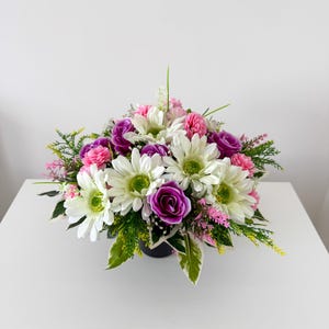 May include: A floral arrangement featuring white daisies with green centres, vibrant purple roses, and small pink blossoms. The bouquet includes various green foliage and sprigs, all contained within a dark, circular base, creating a dense display.