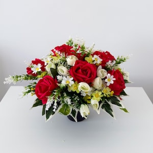 May include: A floral arrangement featuring vibrant red roses, small white flowers, and yellow accents, interspersed with green and white variegated leaves. The bouquet is contained within a dark, rounded base, creating a dense, rounded display.