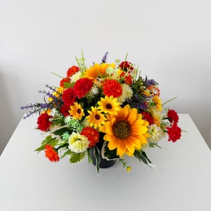 May include: A floral arrangement featuring large yellow sunflowers, vibrant red carnations, and small orange blossoms. The bouquet includes sprigs of purple lavender, white filler flowers, and green leaves, all contained within a dark container.