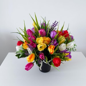 May include: A colourful arrangement of artificial tulips in a black pot. The bouquet features red, yellow, purple, and white tulips, with green grass and lavender accents. The arrangement is set on a white surface.