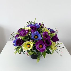 May include: A colourful floral display in a black pot. The arrangement includes purple anemones, blue and purple roses, and yellow flowers with orange centres. Green foliage and small pink accents are also present, set against a white backdrop.