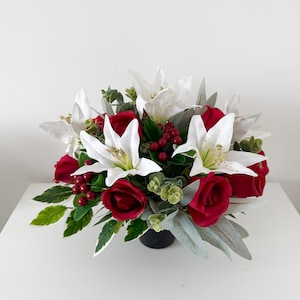 May include: A floral arrangement with red roses and white lilies, complemented by green foliage and red berries. The flowers are presented in a black pot, creating a vibrant display. Ideal for home decoration.