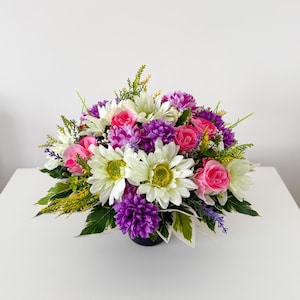 May include: A floral arrangement featuring white daisies with yellow centres, vibrant pink roses, and deep purple pom-pom flowers. The bouquet is accented with green foliage and small sprigs of lavender, all contained within a dark base.