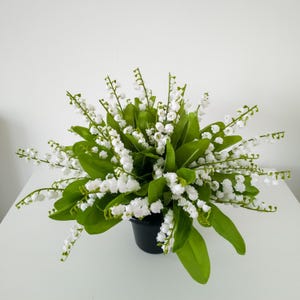 May include: A full arrangement of artificial lily of the valley flowers in a black pot. The bouquet features numerous white bell-shaped blossoms and vibrant green leaves, creating a full and natural appearance. The flowers are arranged to create a visually appealing display.