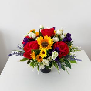 May include: A floral arrangement featuring large red roses, a central yellow sunflower, smaller yellow sunflowers, purple blossoms, and white buds. The bouquet includes green foliage and sprigs of lavender, all contained within a dark container.