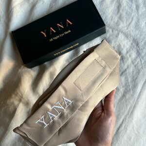 May include: A beige eye mask with the embroidered word "YANA" in white, next to a black box with the word "YANA" and the text "All Night Eye Mask". The website www.yanasleep.com is also visible.