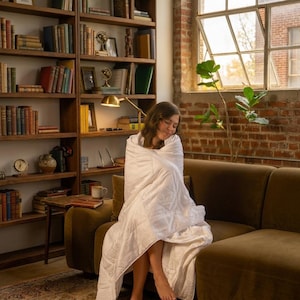 May include: A white weighted blanket with a quilted grid pattern draped over a person sitting on a dark olive green sofa. The blanket features a soft, smooth texture and covers the body, providing a clean, neutral aesthetic in a room.