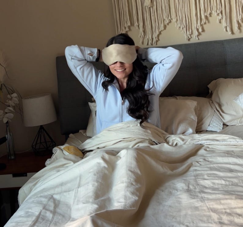 May include: A beige sleep mask is held up by a person wearing a light blue zip-up jacket. The person is sitting on a bed with white bedding. A lamp and decorative wall hanging are in the background.