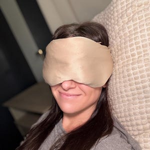 May include: A beige sleep mask covers the eyes. The mask is made of a soft, silky material. The person is wearing a grey long-sleeved top and is resting on a beige pillow.