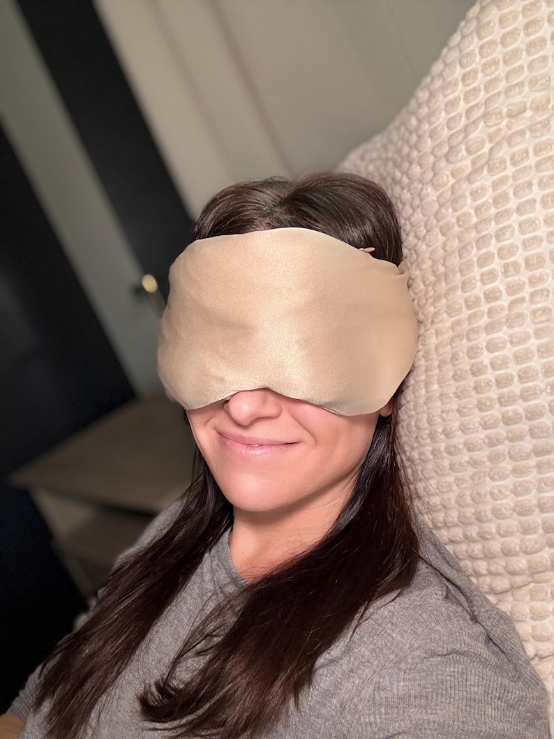 May include: A beige sleep mask covers the eyes. The mask is made of a soft, silky material. The person is wearing a grey long-sleeved top and is resting on a beige pillow.