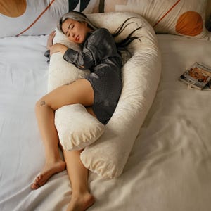 May include: A person sleeping on a white U-shaped body pillow with a white sheet and patterned pillows. The pillow is made of a soft, plush material.