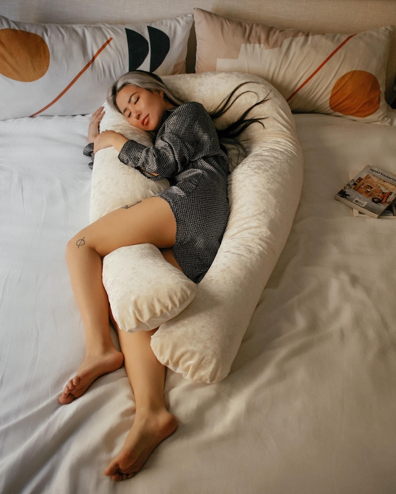 May include: A person sleeping on a white U-shaped body pillow with a white sheet and patterned pillows. The pillow is made of a soft, plush material.