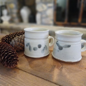 Puede incluir: Dos tazas de cerámica blanca con asas, decoradas con diseños grises pintados a mano. Se encuentran sobre una superficie de madera, junto a una gran piña. El fondo está borroso, sugiriendo un ambiente interior.
