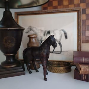 May include: A dark brown horse figurine stands on a white surface, with a framed horse print in the background. A brass bowl and stacked books are also visible. A dark lamp with a metallic shade is on the left.