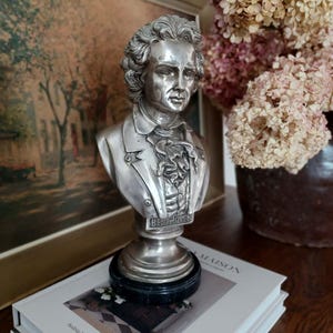 May include: A silver-colored bust of Beethoven, featuring detailed facial features and a ruffled collar. The bust is mounted on a black base and sits atop a stack of books. The word "Beethoven" is inscribed on the bust. A painting and dried flowers are in the background.