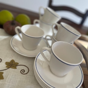 Puede incluir: Un conjunto de tazas y platillos de cerámica blanca con un fino borde azul. Las tazas están apiladas y dispuestas sobre una superficie clara, lo que sugiere una fiesta de té o un comedor. La imagen tiene una iluminación suave y natural.